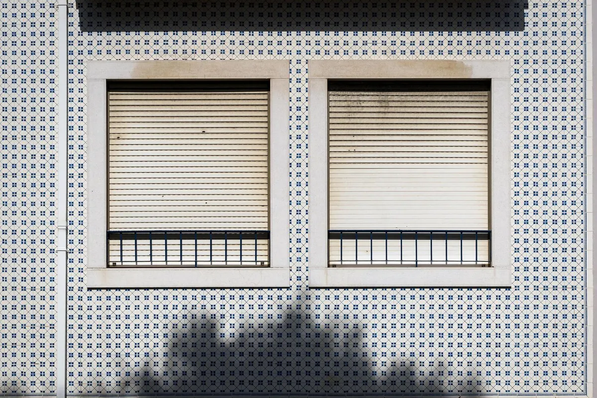Deux volets roulants beiges baissés derrière des garde-corps, sur une façade d'immeuble carrelée blanc et bleu